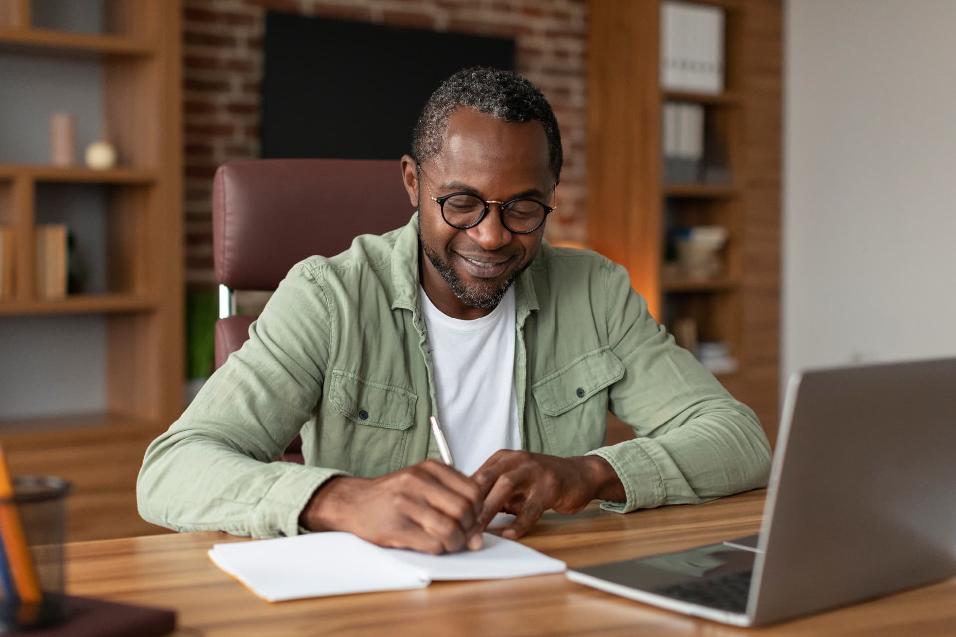 Smiling professional in glasses journaling at his desk beside a laptop