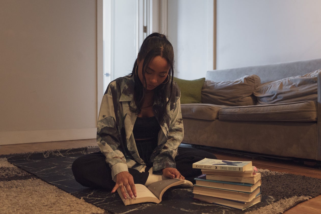 A woman sitting on the floor of a living room reading a book beside a stack of books